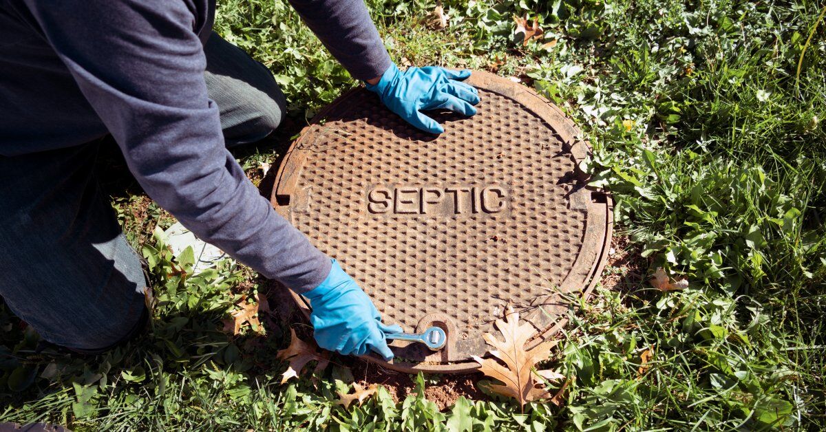 A person wearing blue gloves uses a small wrench to open the entrance to a septic system. The cover has the word "SEPTIC" on it.