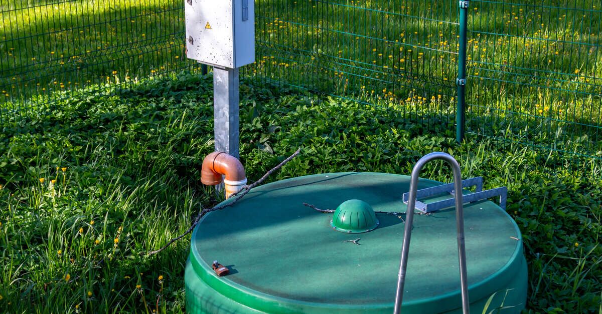 A bright green septic tank peeks out over grass and weeds in an area that is fenced off on a sunny day.