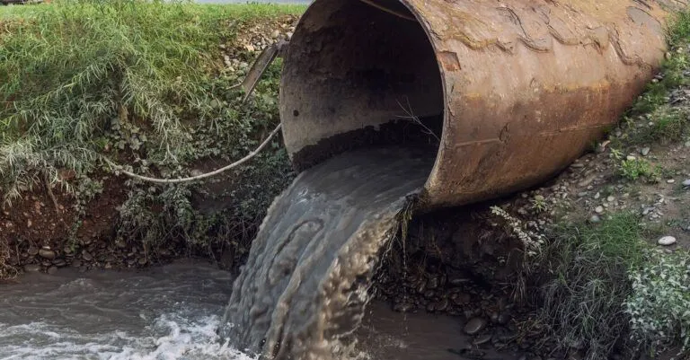 A close-up showing a large, brown pipe coming out of the ground that is emitting dark colored sewage.