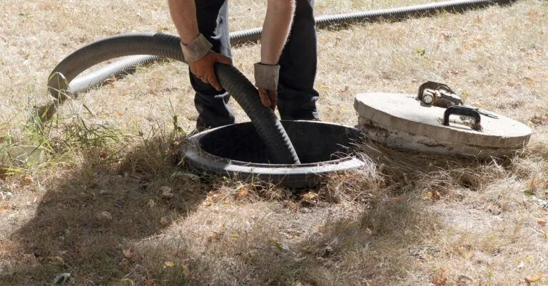 A person wearing a yellow high-visibility vest and orange gloves inserts a large pipe into a septic tank.