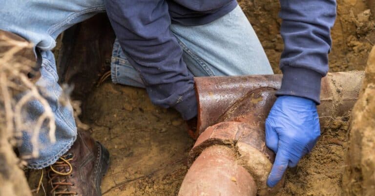 A man wearing work gloves and boots trying to clear away dirt and debris from a severely blocked sewer line.