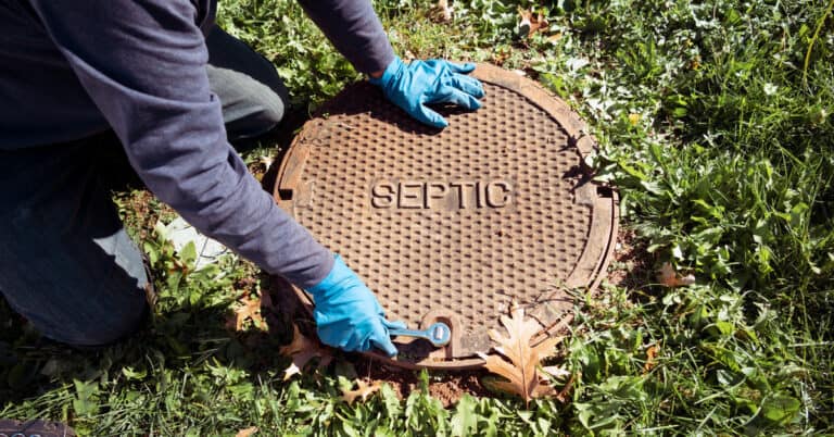 A working plumber opens a septic system tank in the ground for maintenance while wearing gloves on a sunny day.