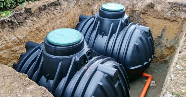 Two large black plastic septic tanks attached to an orange pipe are in an underground hole next to a bush.