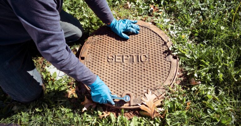 A person wearing blue gloves uses a small wrench to open the entrance to a septic system. The cover has the word "SEPTIC" on it.