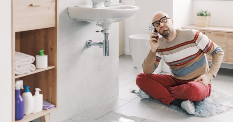 A bearded man in glasses is talking on a cellphone while sitting on a bathroom floor in front of a puddle of water.