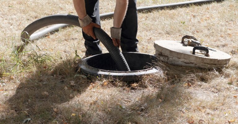 A person wearing a yellow high-visibility vest and orange gloves inserts a large pipe into a septic tank.