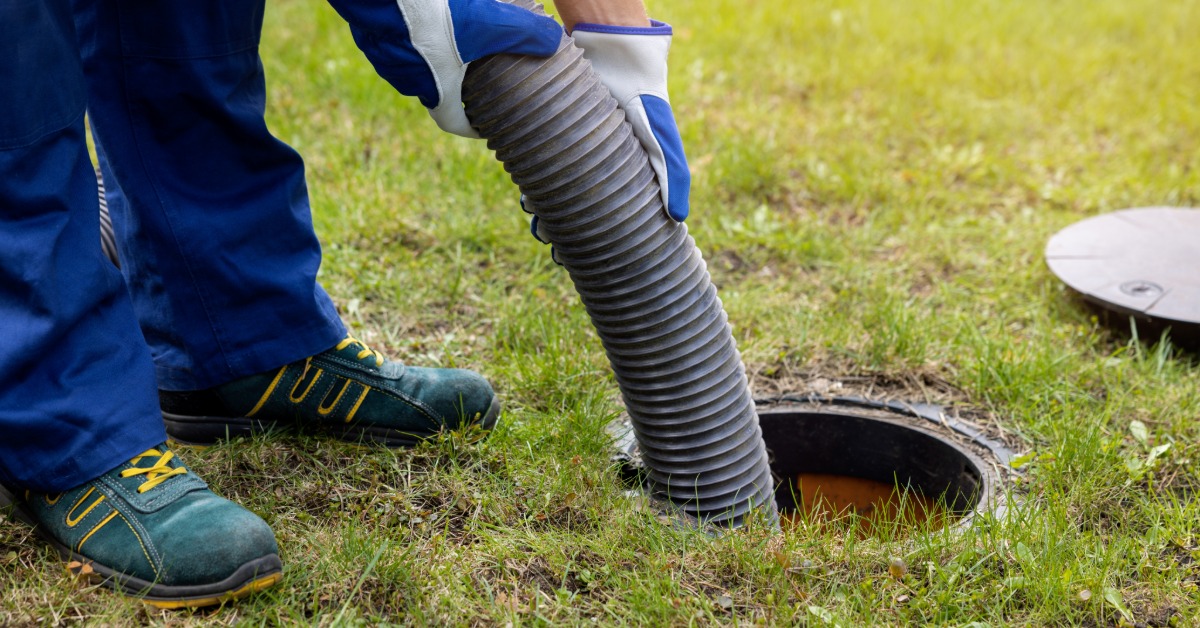 A plumber holding a hose into a septic system tank to pump out the contents. The opening to the tank is underground.