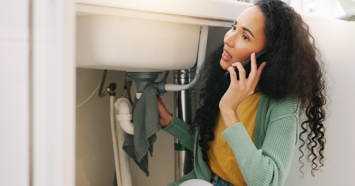 A woman on the phone while holding a towel up to the pipes under the sink. There is a plumbing problem with the pipes.