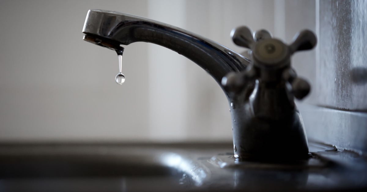 A clean bathroom faucet with a knob handle is leaking, with a small droplet of water falling from the spout.