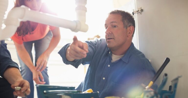 A man pointing beneath a sink while another person works with tools, and a woman leaning nearby in a cabinet.