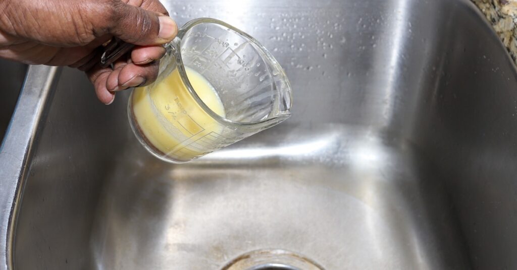 A person holding a measuring cup with old cooking grease over a kitchen sink, ready to pour it down the drain.