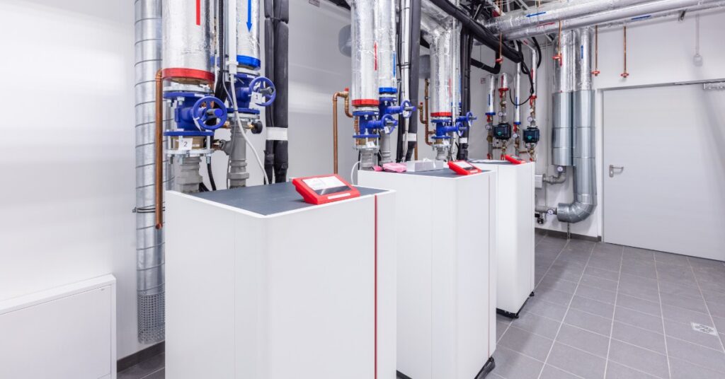 A bright mechanical room with white heat pump units, silver insulated pipes, blue valves, gauges, and gray tile floor.