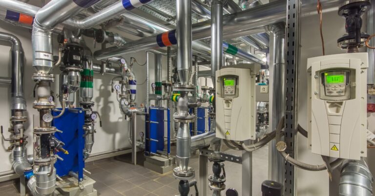 A boiler room with red pipes, valves, gauges, pumps, metal tanks, and insulated lines mounted on a wall.