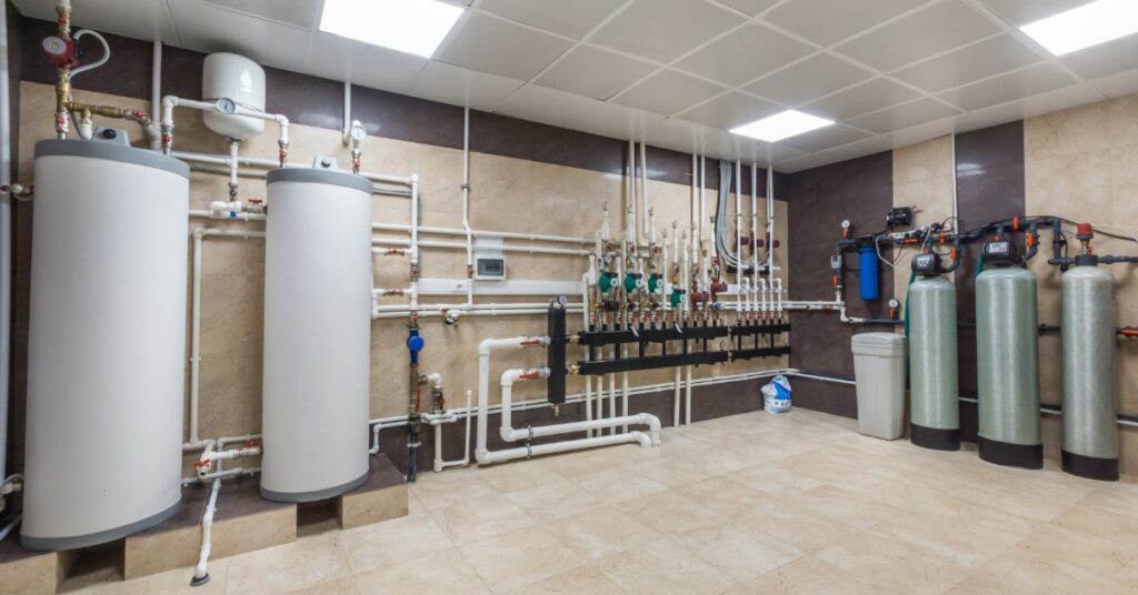 A utility room with two water heaters, silver pipes, valves, pressure gauges, and expansion tanks along white walls.