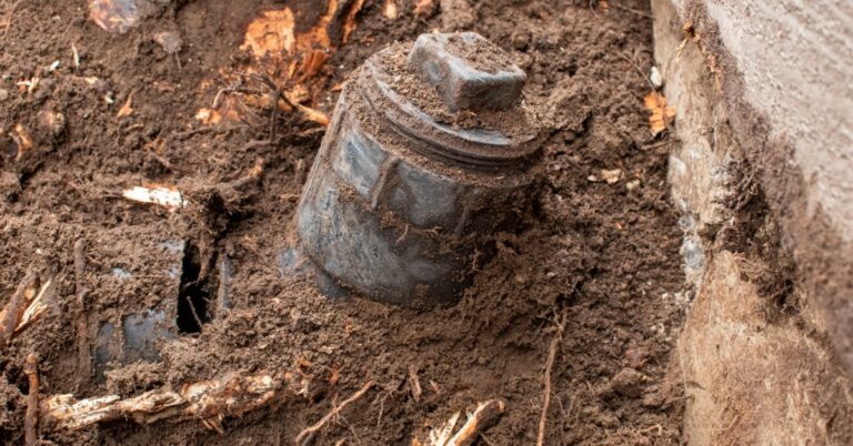 A damaged septic pipe cap partially buried in dark soil beside tree roots and a concrete foundation wall.