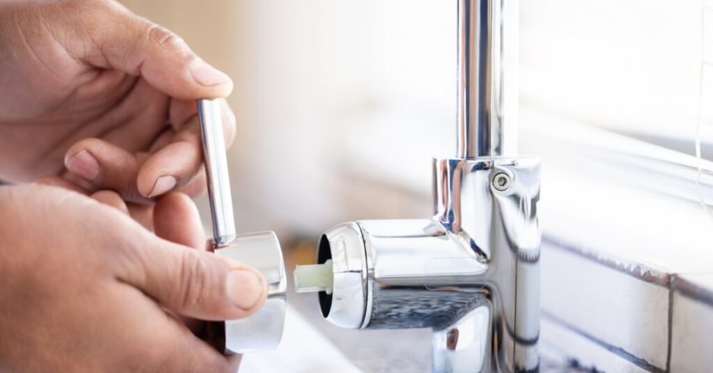 A pair of hands attaching a chrome faucet handle beside a shiny tap, white tile, and a bright window.