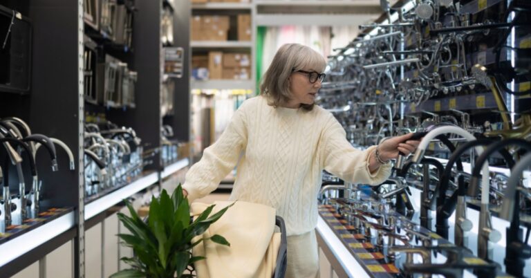 A woman in a cream sweater reaching for a faucet display while pushing a cart in a hardware store aisle.
