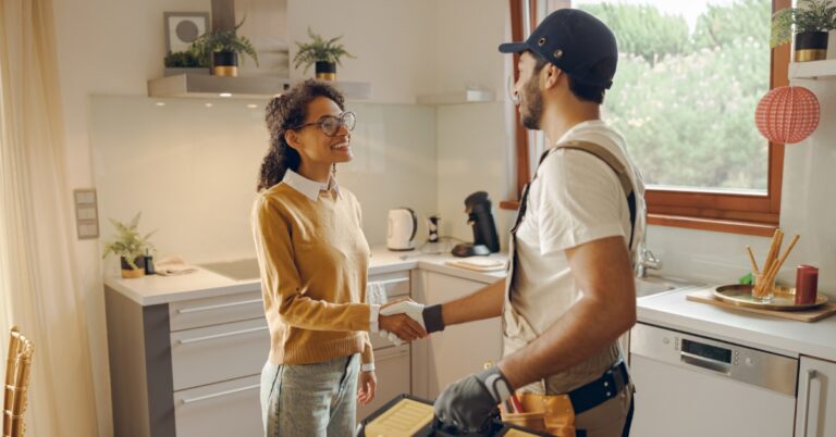 A repairman in a dark uniform shaking hands with a woman in a kitchen, with white cabinets and a toolbox nearby.