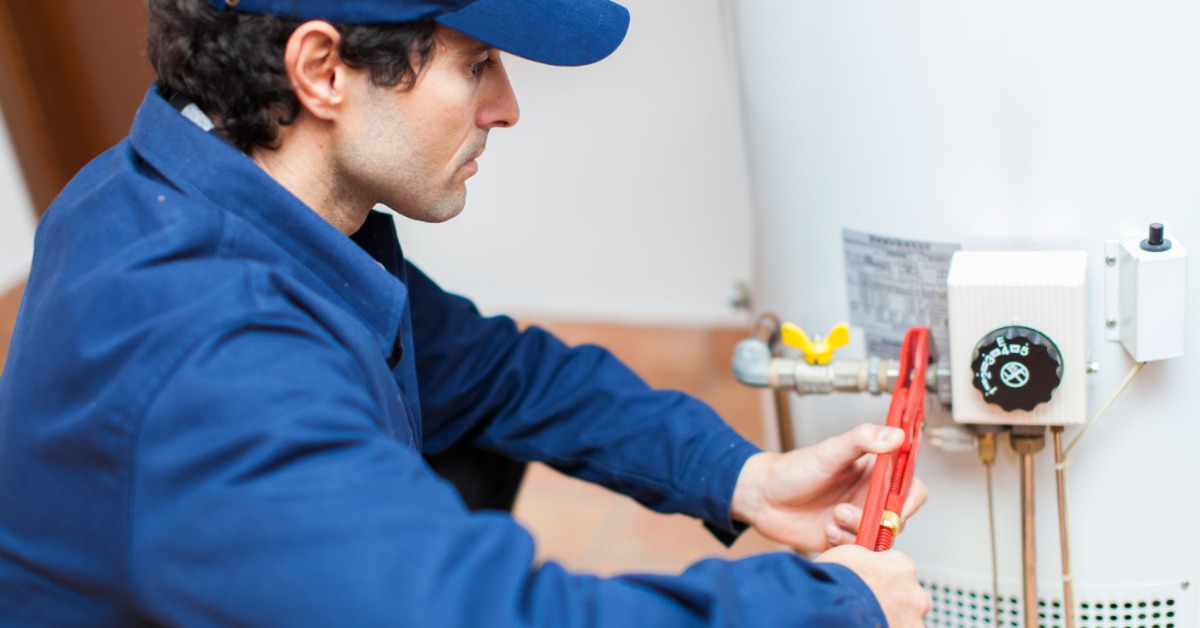 A plumber kneeling beside an open white water heater while using tools near copper pipes and gray wall tiles.