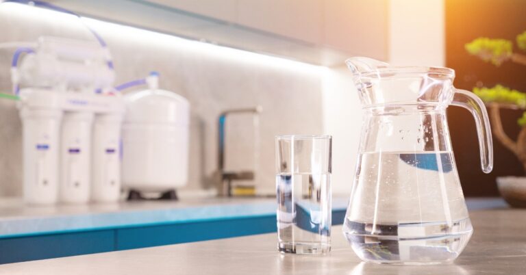 A glass pitcher and glass of water on a counter, with a blurred water filter system and kitchen sink behind them.
